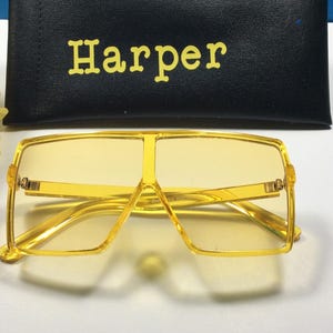 May include: Yellow square-shaped sunglasses with a gold frame. The sunglasses are sitting on a white surface. The brand name "Harper" is printed on a black case.