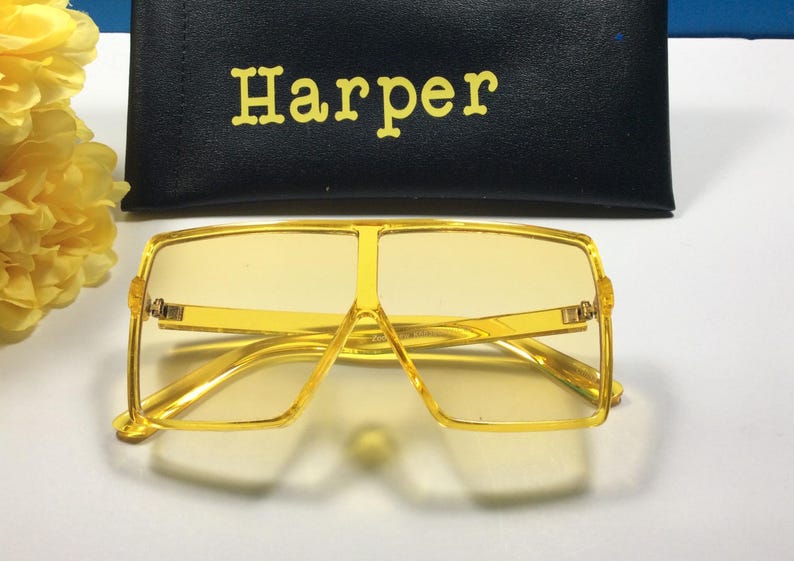 May include: Yellow square-shaped sunglasses with a gold frame. The sunglasses are sitting on a white surface. The brand name "Harper" is printed on a black case.