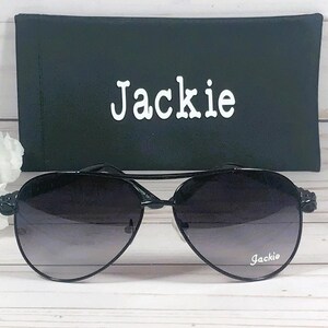Peut inclure: Lunettes de soleil noires avec des verres d&eacute;grad&eacute;s et un &eacute;tui noir. L'&eacute;tui porte le nom "Jackie" imprim&eacute; en blanc.