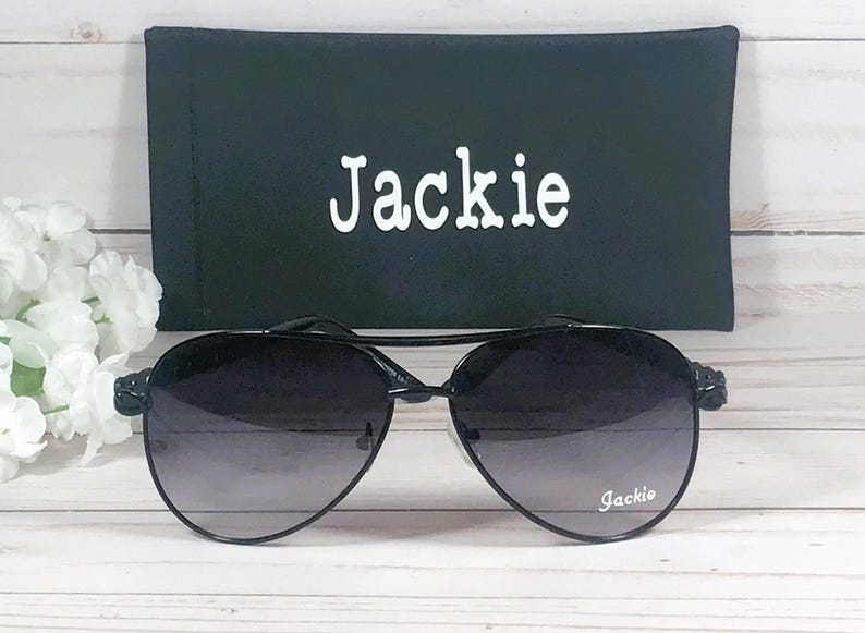 Peut inclure: Lunettes de soleil noires avec des verres d&eacute;grad&eacute;s et un &eacute;tui noir. L'&eacute;tui porte le nom "Jackie" imprim&eacute; en blanc.