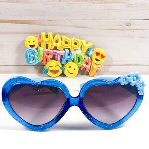 May include: Blue heart-shaped sunglasses with blue flower accents. The sunglasses have a dark lens. The sunglasses are in front of a sign that says "Happy Birthday" with colorful emoji faces.