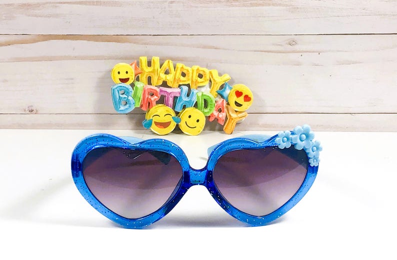 May include: Blue heart-shaped sunglasses with blue flower accents. The sunglasses have a dark lens. The sunglasses are in front of a sign that says "Happy Birthday" with colorful emoji faces.