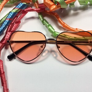May include: Heart-shaped sunglasses with a rose gold frame and pink lenses. The sunglasses have "UV Protection" printed on the right lens.