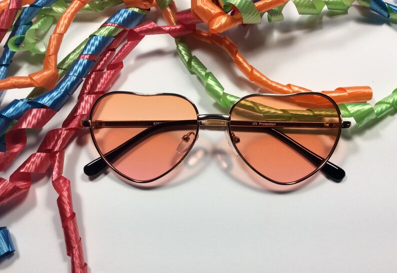May include: Heart-shaped sunglasses with a rose gold frame and pink lenses. The sunglasses have "UV Protection" printed on the right lens.