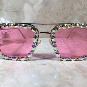 Peut inclure: Lunettes de soleil carr&eacute;es dor&eacute;es avec des verres roses et des strass iris&eacute;s autour des montures.
