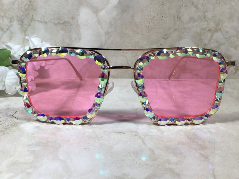 Peut inclure: Lunettes de soleil carr&eacute;es dor&eacute;es avec des verres roses et des strass iris&eacute;s autour des montures.