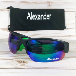 May include: Black sunglasses with bright green inner temples and iridescent purple-to-green reflective lenses featuring the name Alexander printed in white. A matching black drawstring pouch with the name Alexander in white is positioned behind.