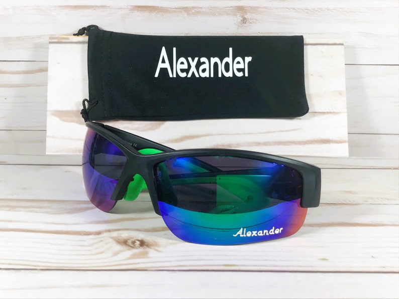 May include: Black sunglasses with bright green inner temples and iridescent purple-to-green reflective lenses featuring the name Alexander printed in white. A matching black drawstring pouch with the name Alexander in white is positioned behind.