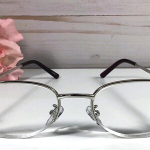 May include: Silver-framed eyeglasses with oval lenses. The frames have dark burgundy temples and are embellished with pink gemstones at the outer edges. A pink flower is in the background.