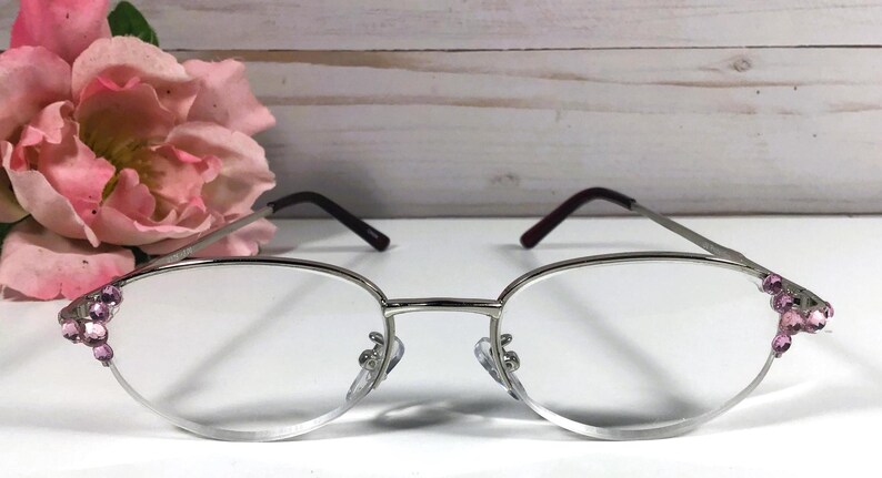 May include: Silver-framed eyeglasses with oval lenses. The frames have dark burgundy temples and are embellished with pink gemstones at the outer edges. A pink flower is in the background.