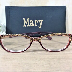 May include: Burgundy reading glasses with gold embellishments along the top rim. The glasses are paired with a black case with the name "Mary" in gold lettering. The glasses have a +2.00 magnification.