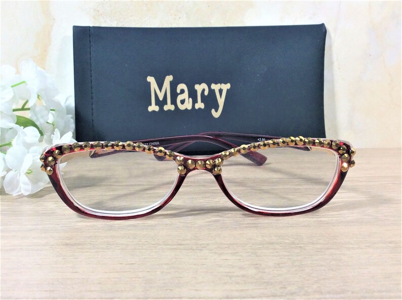 May include: Burgundy reading glasses with gold embellishments along the top rim. The glasses are paired with a black case with the name "Mary" in gold lettering. The glasses have a +2.00 magnification.