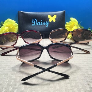 Op de afbeelding: Drie zonnebrillen met gouden accenten en verschillende gekleurde monturen. De zonnebrillen liggen op een gepatroneerd oppervlak met een zwarte etui erachter. De etui heeft het woord "Daisy" en een vlinderontwerp erop.