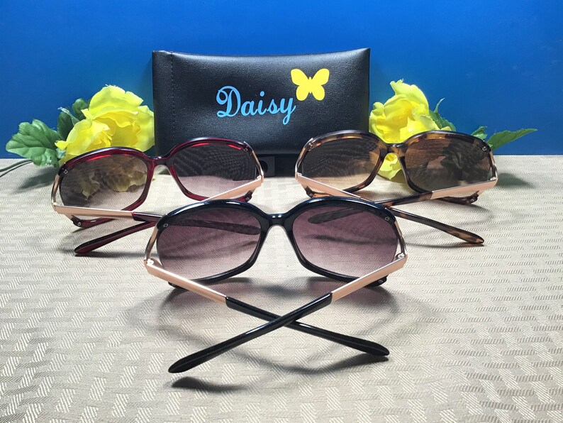 Op de afbeelding: Drie zonnebrillen met gouden accenten en verschillende gekleurde monturen. De zonnebrillen liggen op een gepatroneerd oppervlak met een zwarte etui erachter. De etui heeft het woord "Daisy" en een vlinderontwerp erop.