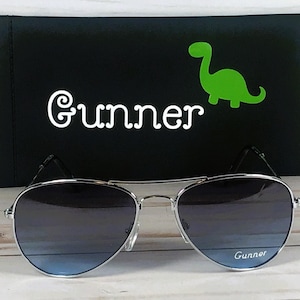 May include: Silver-framed aviator sunglasses with gradient blue lenses featuring the word Gunner printed on the right lens. Behind the sunglasses is a black rectangular case with the name Gunner and a bright green dinosaur graphic.