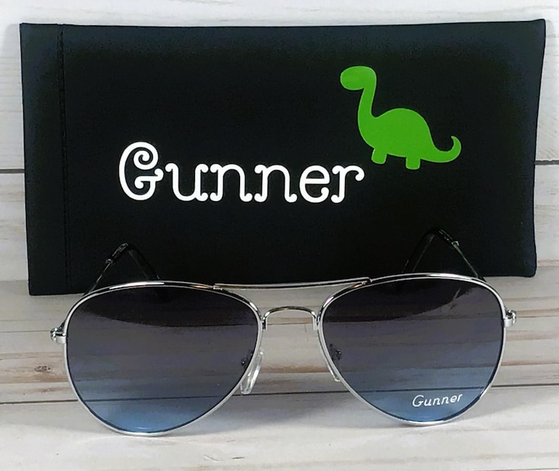 May include: Silver-framed aviator sunglasses with gradient blue lenses featuring the word Gunner printed on the right lens. Behind the sunglasses is a black rectangular case with the name Gunner and a bright green dinosaur graphic.