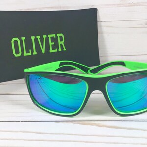 K&ouml;nnte beinhalten: Schwarze Sonnenbrille mit gr&uuml;nen Rahmen und verspiegelten Gl&auml;sern. Die Sonnenbrille befindet sich in einem schwarzen Etui mit dem Namen "OLIVER" in gr&uuml;ner Schrift.
