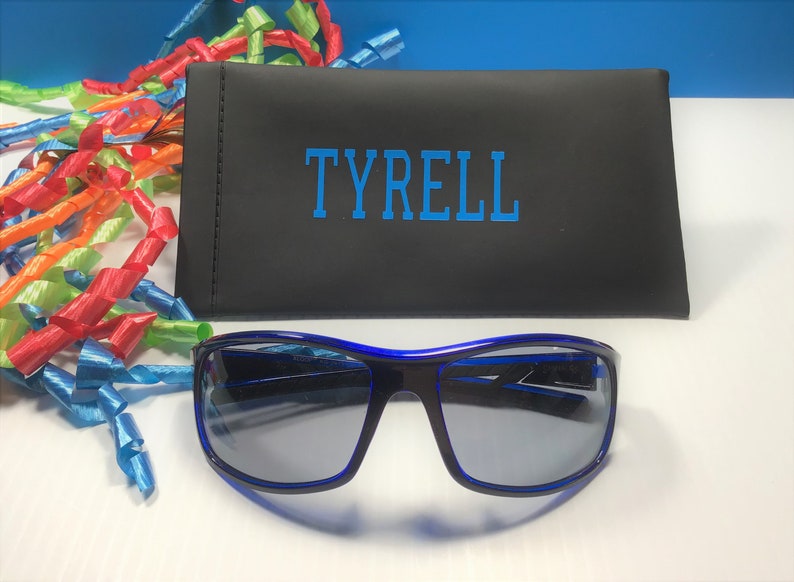 K&ouml;nnte beinhalten: Blaue Sonnenbrille mit schwarzem Rahmen und blauen Gl&auml;sern in einem schwarzen Etui mit dem Namen "TYRELL" in blau.