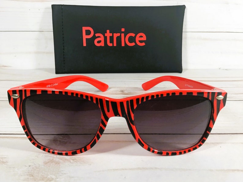 May include: Red and black zebra print sunglasses with dark gray lenses. The sunglasses are in a black case with the word "Patrice" printed in red.