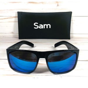 Puede incluir: Gafas de sol negras con lentes espejadas azules y estuche negro. El estuche tiene el nombre "Sam" impreso en blanco. Las gafas tienen una montura gruesa y cuadrada.
