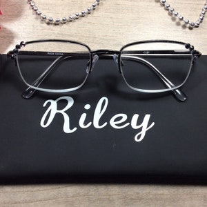 May include: Black-framed eyeglasses with clear lenses rest on a black case. The case is labeled "Riley" in white script. The glasses have small decorative accents on the temples. A silver beaded necklace and a red flower are in the background.