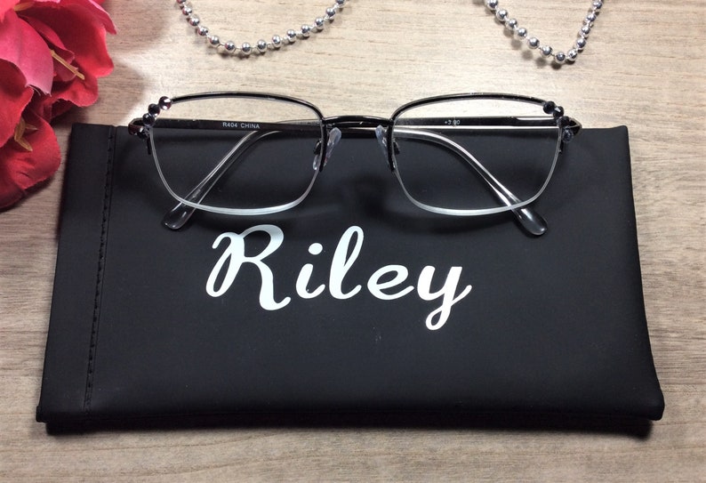 May include: Black-framed eyeglasses with clear lenses rest on a black case. The case is labeled "Riley" in white script. The glasses have small decorative accents on the temples. A silver beaded necklace and a red flower are in the background.