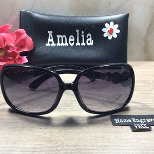 Puede incluir: Gafas de sol negras con un dise&ntilde;o en forma de coraz&oacute;n en las patillas. Las gafas de sol vienen con una funda negra con el nombre "Amelia" grabado y una margarita blanca con un centro rojo.