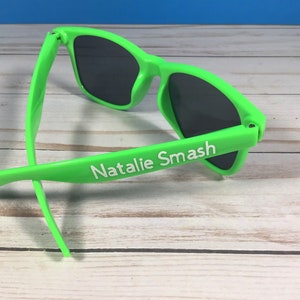 Op de afbeelding: Groene zonnebril met zwarte glazen en de naam "Natalie Smash" in wit op de zijkant.