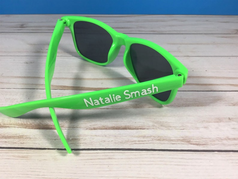 Op de afbeelding: Groene zonnebril met zwarte glazen en de naam "Natalie Smash" in wit op de zijkant.