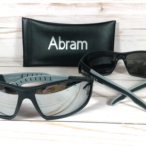 May include: A pair of black sunglasses with mirrored lenses and gray accents. The sunglasses are in a black case with the name "Abram" printed on it.