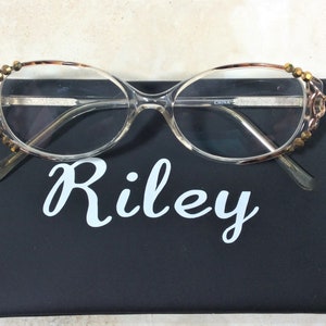Puede incluir: Gafas con montura transparente y adornos dorados en las patillas sobre un estuche negro. El estuche tiene el nombre "Riley" en escritura blanca. Las gafas tienen una montura transparente y est&aacute;n dise&ntilde;adas para la correcci&oacute;n de la visi&oacute;n.