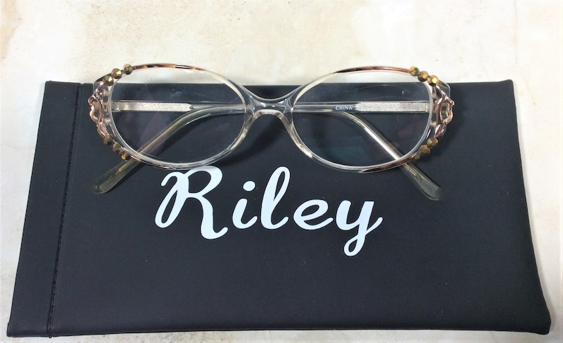 Puede incluir: Gafas con montura transparente y adornos dorados en las patillas sobre un estuche negro. El estuche tiene el nombre "Riley" en escritura blanca. Las gafas tienen una montura transparente y est&aacute;n dise&ntilde;adas para la correcci&oacute;n de la visi&oacute;n.
