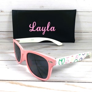 Op de afbeelding: Roze zonnebril met een wit montuur en een vredesduif-patroon op de pootjes. De zonnebril wordt geleverd met een zwarte etui met de naam "Layla" erop geborduurd in roze.