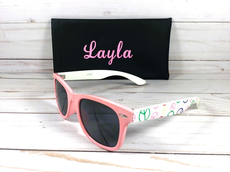 Op de afbeelding: Roze zonnebril met een wit montuur en een vredesduif-patroon op de pootjes. De zonnebril wordt geleverd met een zwarte etui met de naam "Layla" erop geborduurd in roze.