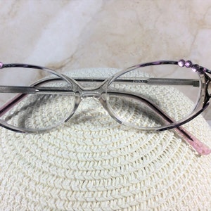 Puede incluir: Gafas de forma ovalada con montura transparente y detalles morados. La parte superior de la montura est&aacute; adornada con peque&ntilde;os brillantes morados. Las patillas son de color rosa claro. Las gafas est&aacute;n sobre un sombrero blanco tejido.