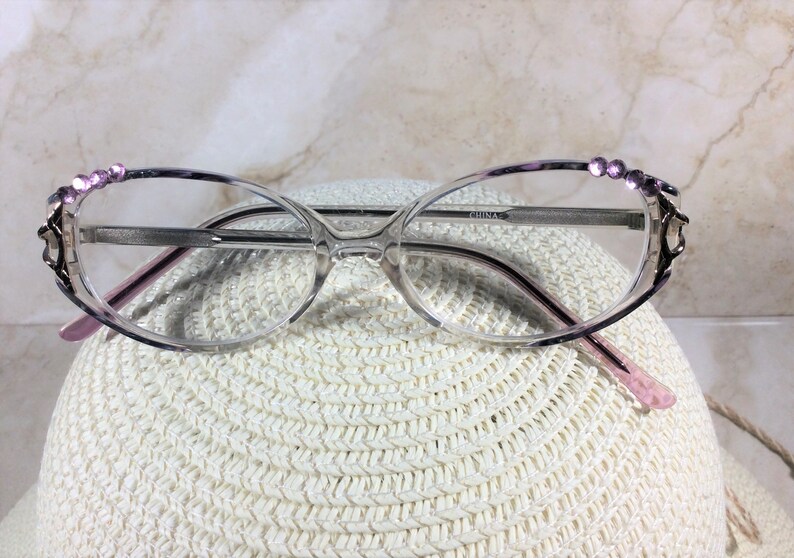Puede incluir: Gafas de forma ovalada con montura transparente y detalles morados. La parte superior de la montura est&aacute; adornada con peque&ntilde;os brillantes morados. Las patillas son de color rosa claro. Las gafas est&aacute;n sobre un sombrero blanco tejido.