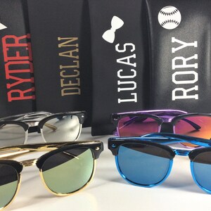 May include: Four personalized sunglass cases with different designs, including a star, a bow tie, a baseball, and the names "Ryder", "Declan", "Lucas", and "Rory".  The cases are black with white lettering.  Four pairs of sunglasses with different colored lenses are shown in front of the cases.
