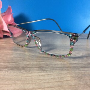 May include: Clear eyeglasses with a silver frame and colorful rhinestones along the rims. The rhinestones are in shades of pink, green, and blue. The glasses are set against a blue background.