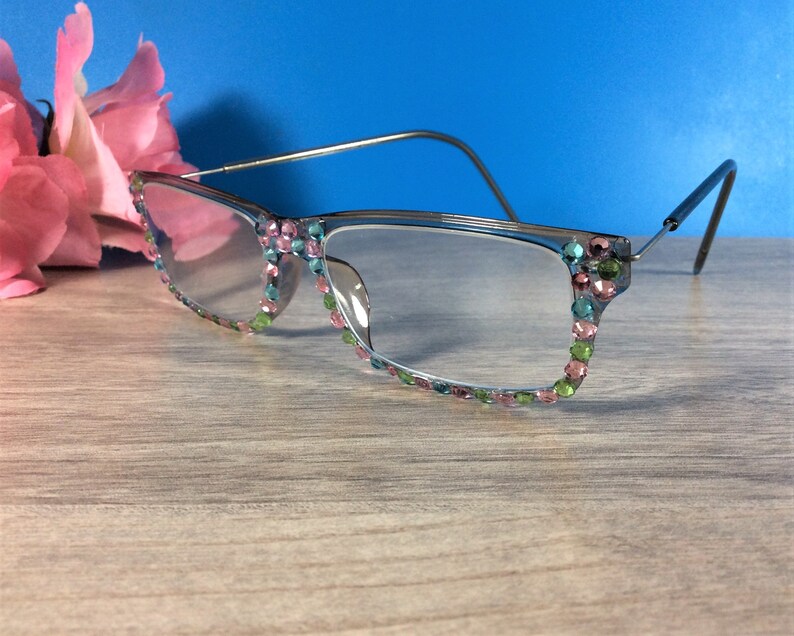 May include: Clear eyeglasses with a silver frame and colorful rhinestones along the rims. The rhinestones are in shades of pink, green, and blue. The glasses are set against a blue background.