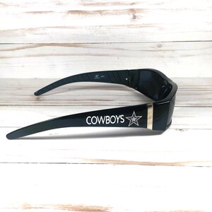 May include: Black sunglasses with the word "COWBOYS" and a star logo printed in white. The sunglasses have a gold-colored stripe on the side and are set against a light wood background.