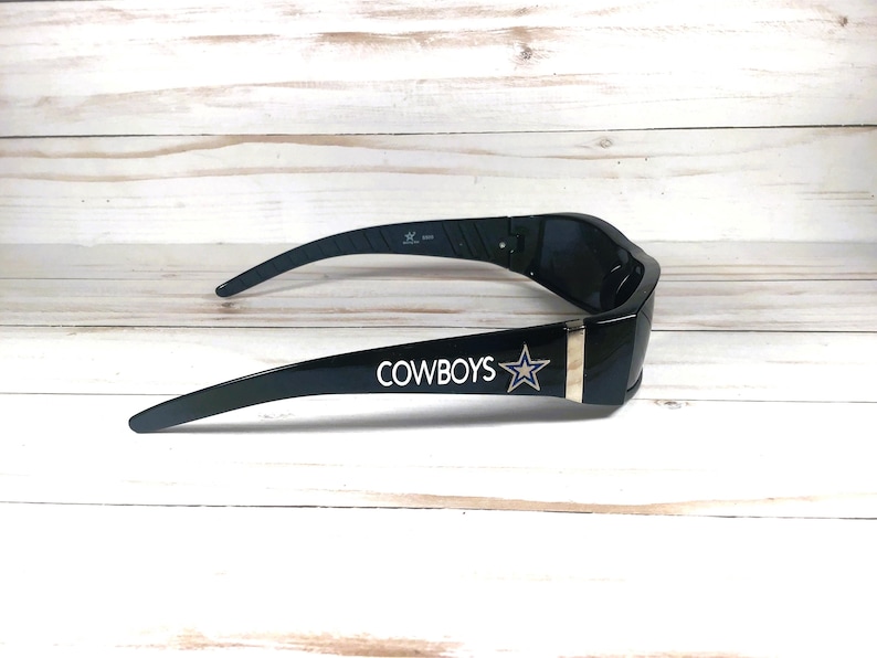 May include: Black sunglasses with the word "COWBOYS" and a star logo printed in white. The sunglasses have a gold-colored stripe on the side and are set against a light wood background.