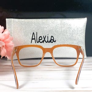 Op de afbeelding: Oranje montuurbril met bijpassende etui. De etui is wit met zwarte spikkels en de naam "Alexis" in zwart schrift. De bril staat tegen een vage achtergrond met roze bloemen.