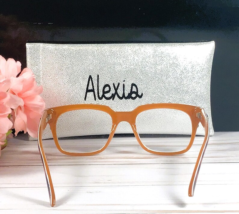 Op de afbeelding: Oranje montuurbril met bijpassende etui. De etui is wit met zwarte spikkels en de naam "Alexis" in zwart schrift. De bril staat tegen een vage achtergrond met roze bloemen.