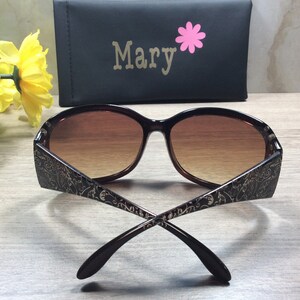 K&ouml;nnte beinhalten: Braune Sonnenbrille mit goldenem Blumenmuster an den B&uuml;geln. Die Sonnenbrille ist zusammengeklappt und die B&uuml;gel sind gekreuzt.