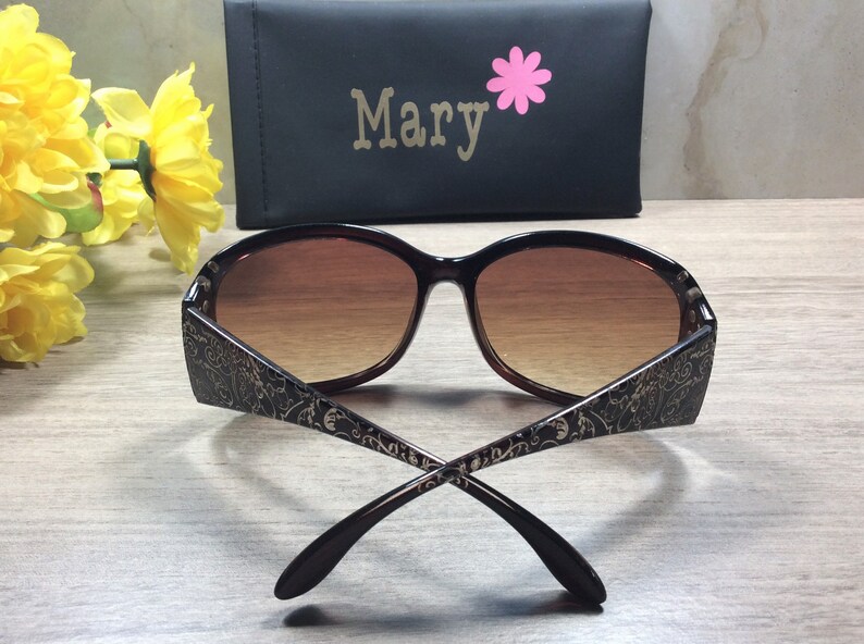 K&ouml;nnte beinhalten: Braune Sonnenbrille mit goldenem Blumenmuster an den B&uuml;geln. Die Sonnenbrille ist zusammengeklappt und die B&uuml;gel sind gekreuzt.