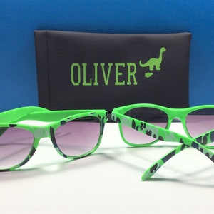 Może przedstawiać: Zielone okulary przeciwsłoneczne z wzorem kamuflażu i czarnym etui z napisem "OLIVER" i zielonym dinozaurem.