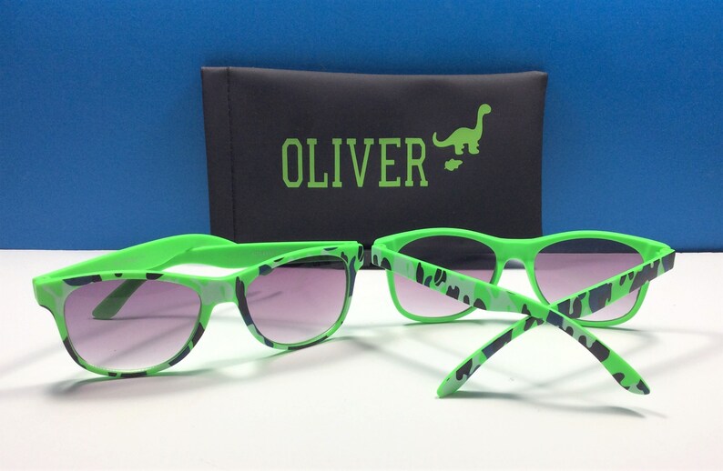 Może przedstawiać: Zielone okulary przeciwsłoneczne z wzorem kamuflażu i czarnym etui z napisem "OLIVER" i zielonym dinozaurem.