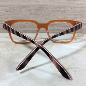 Puede incluir: Gafas con montura naranja y varillas a rayas marrones y blancas. Las gafas tienen forma cuadrada y est&aacute;n sobre un fondo claro. Las varillas se cruzan.