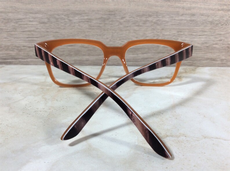 Puede incluir: Gafas con montura naranja y varillas a rayas marrones y blancas. Las gafas tienen forma cuadrada y est&aacute;n sobre un fondo claro. Las varillas se cruzan.