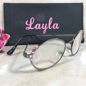 Puede incluir: Gafas con montura plateada, lentes transparentes y adornos de joyas rosas. Las gafas est&aacute;n frente a un estuche negro con el nombre "Layla" en escritura rosa. Una flor rosa est&aacute; en el fondo.